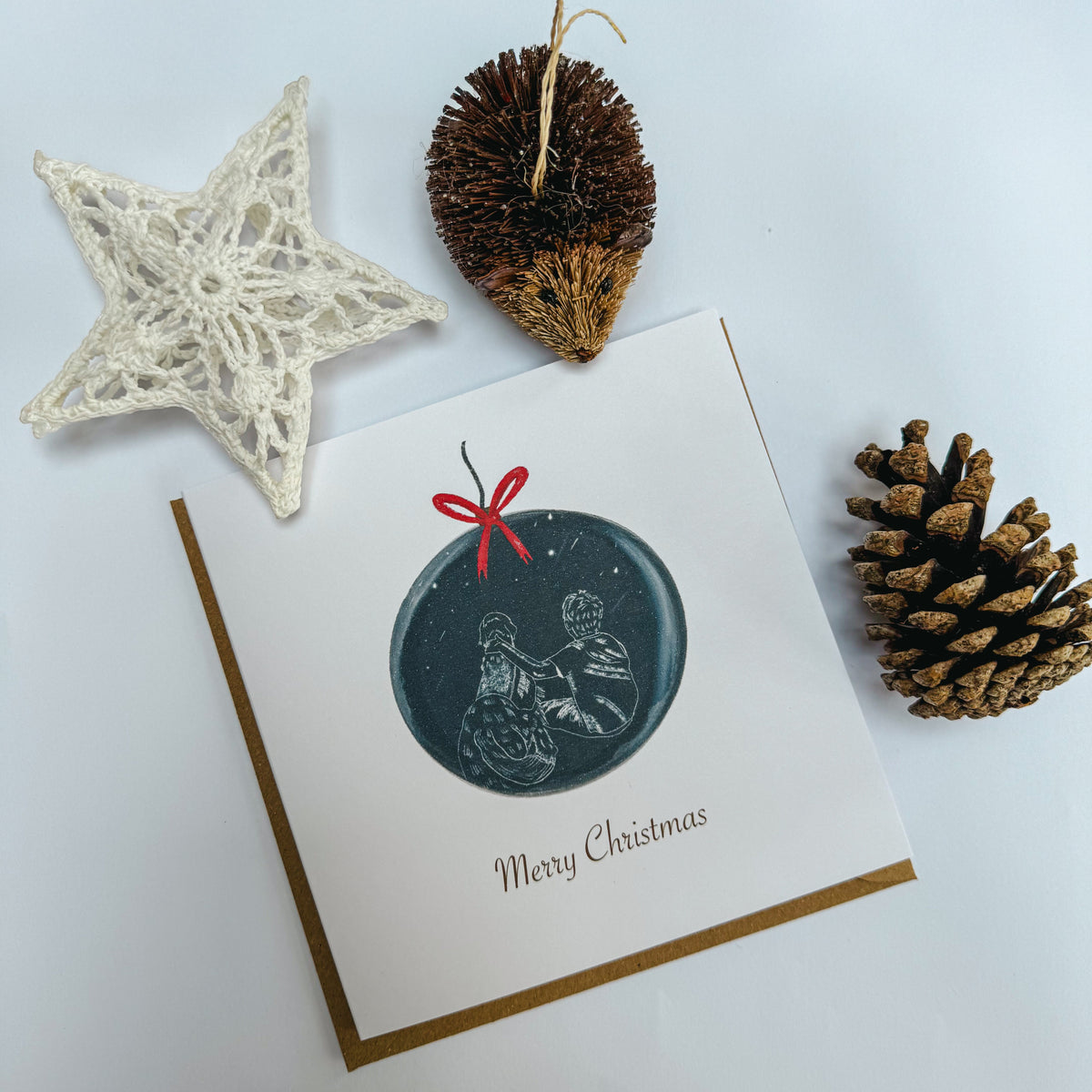 A square white Christmas greeting card featuring a circular dark blue bauble illustration with a red bow and the words 'Merry Christmas' below it. On the bauble is a sketch of an assistance dog and their handler. The card is styled on a white background with a cream lace star and a pinecone.