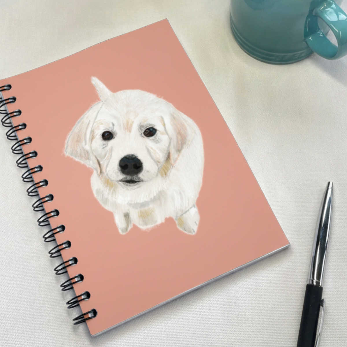 A spiral-bound notebook with a dusty pink cover featuring a painted white puppy illustration, styled on a light fabric surface with a teal mug and a black pen nearby.