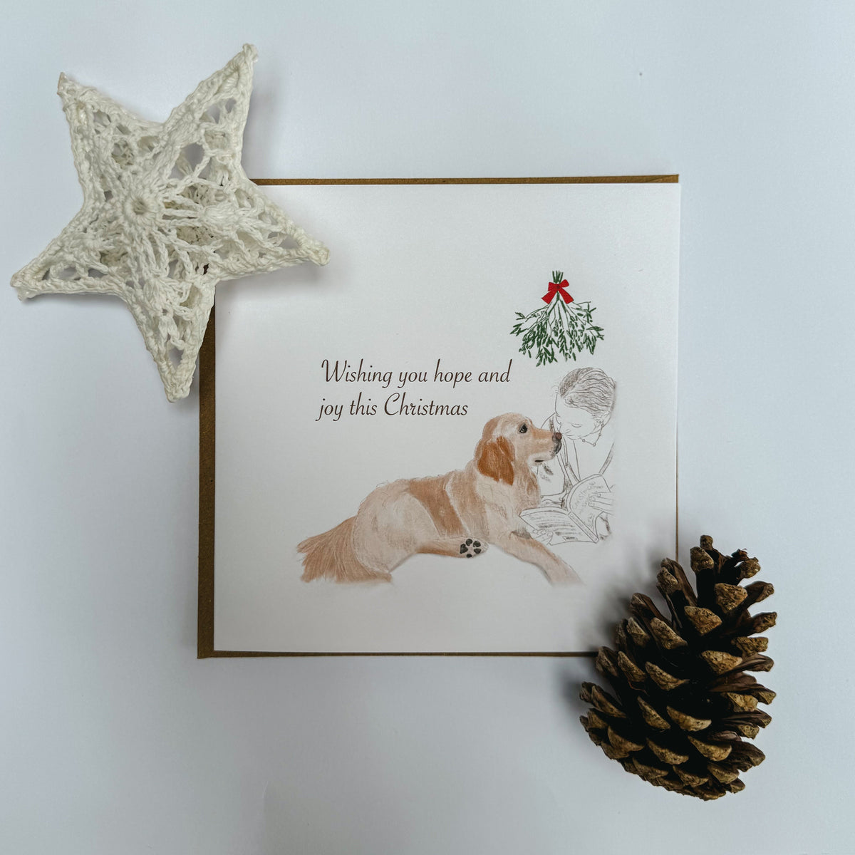 A square Christmas greeting card featuring an illustration of a golden retriever and a person with the text 'Wishing you hope and joy this Christmas' and a small mistletoe graphic. The card is styled on a white background with a white star ornament and a pinecone.