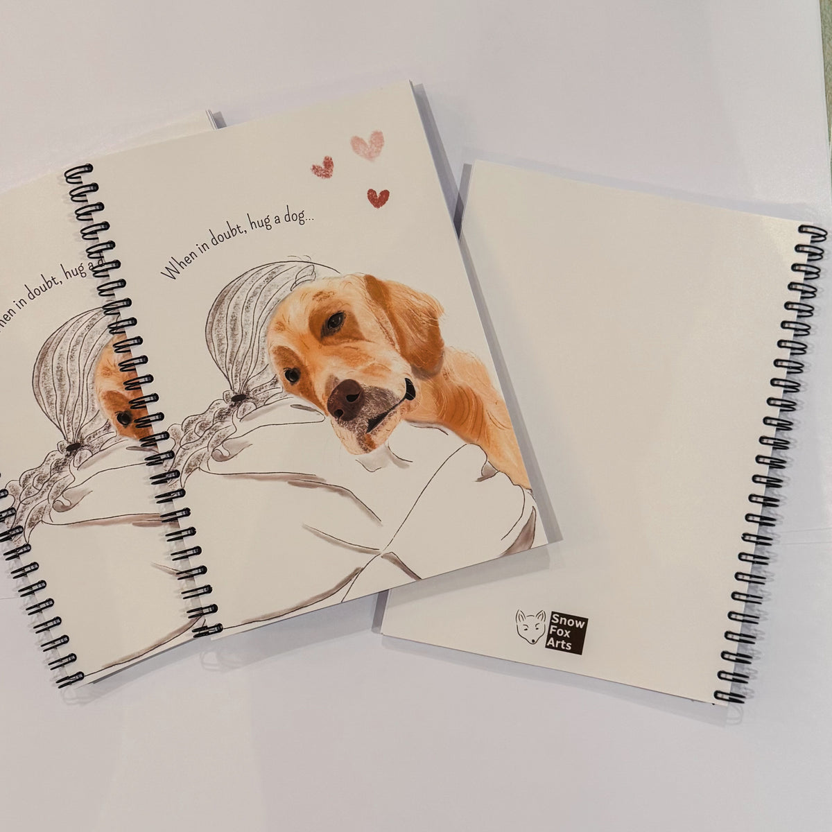 Three spiral-bound notebooks on a white surface. The top notebook features a watercolor illustration of a golden retriever dog hugging a woman, with the text 'When a doubt, hug a dog' and small hearts.