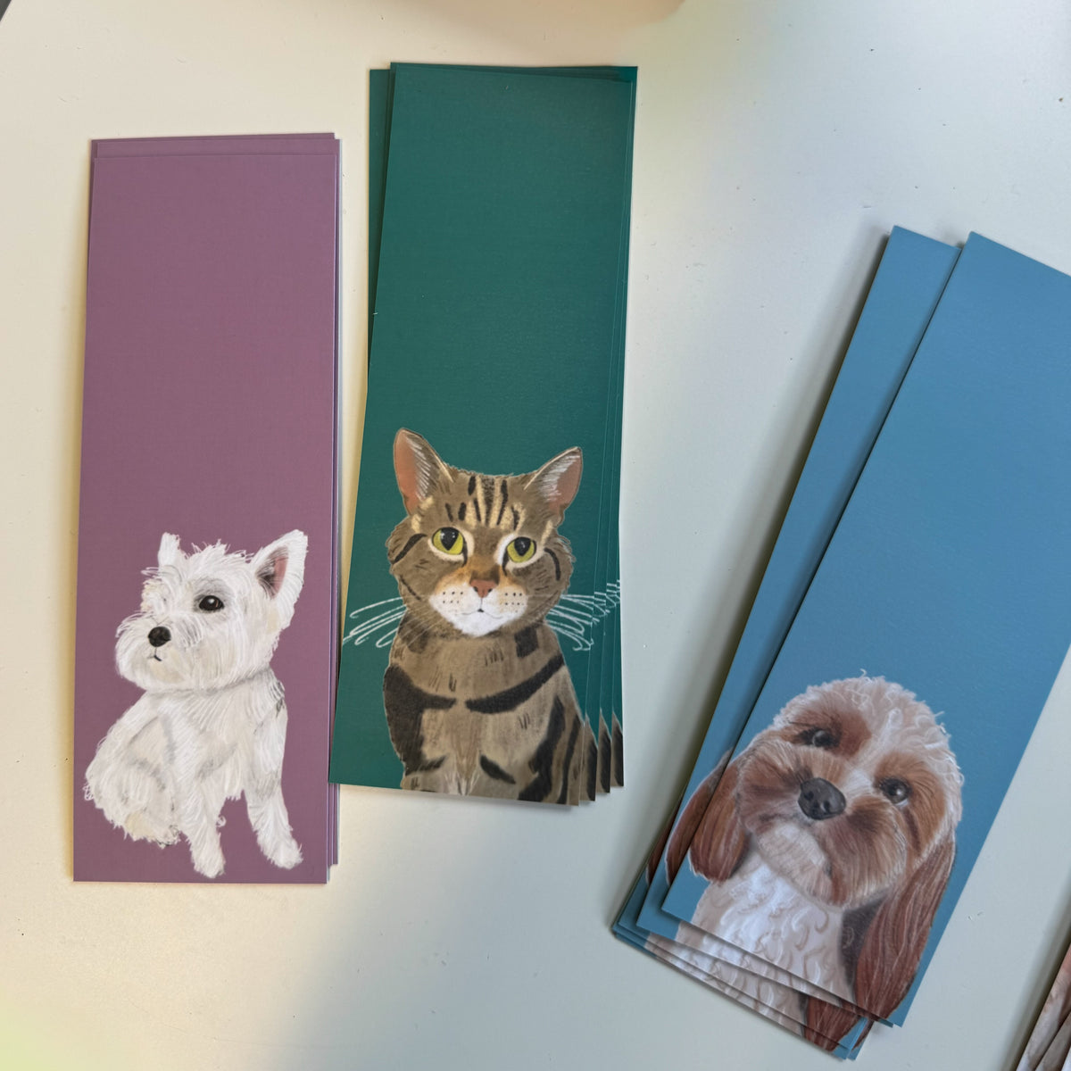 Three bookmarks with painted pet illustrations are arranged on a light surface. One card shows a white dog on a mauve background, one shows a brown tabby cat on a dark green background, and a stack of blue cards on the right shows a brown-and-white dog portrait.