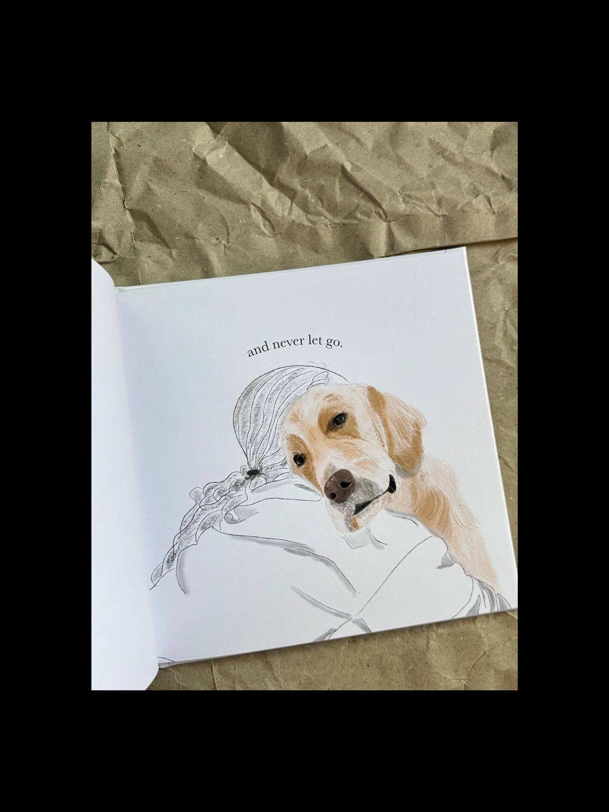 An open book showing a painted illustration of a golden retriever resting on a white page with the text 'and never let go.' The page is placed on crumpled brown paper.