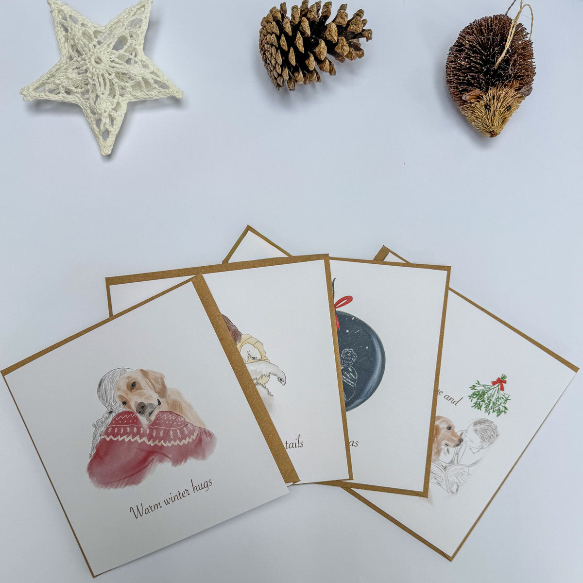 Four Christmas Cards displayed on a white table with a pinecone, a hedgehog ornament and a lace star.