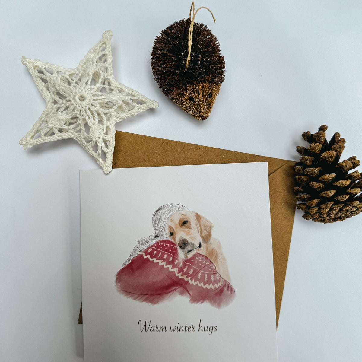 A square greeting card with an illustration of a golden retriever hugging a person in a red knitted sweater. The card is styled on a white background with a white crocheted star, a brown hedgehog ornament, and a pinecone.